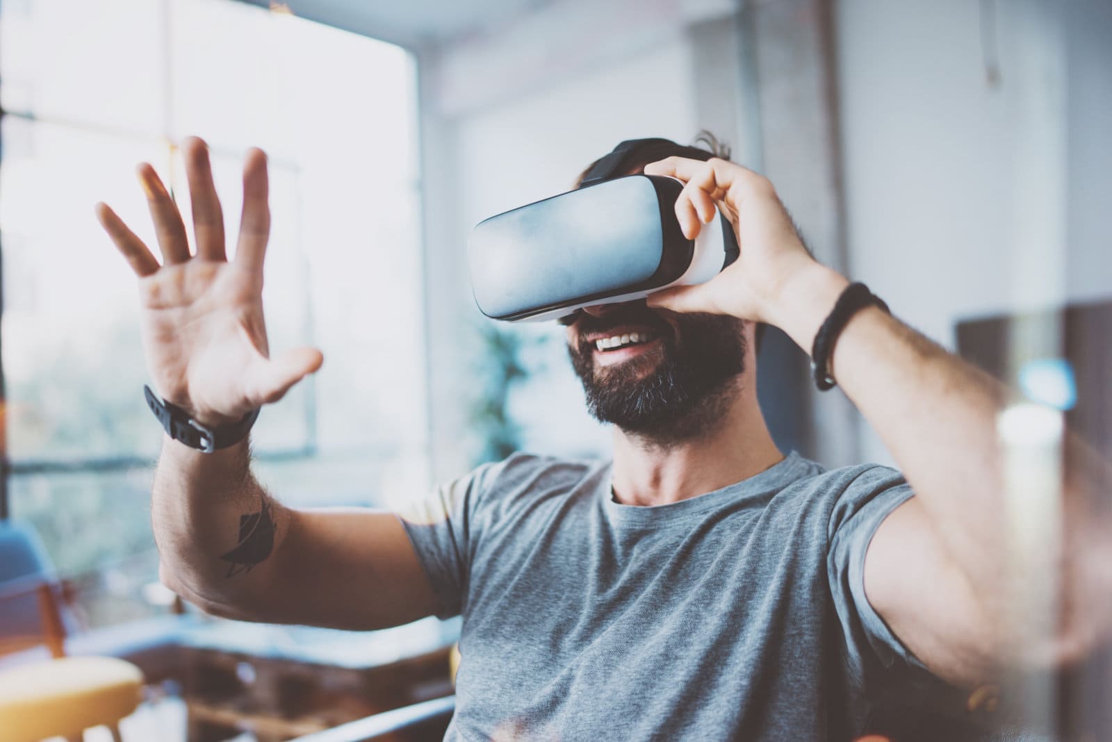 Young bearded man wearing virtual reality glasses in modern interior design coworking studio. Smartphone using with VR goggles headset. Horizontal,flares effect, blurred background.