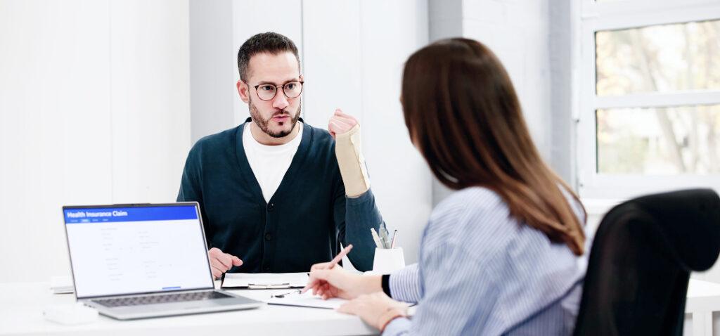 Man with a cast on his arm explains something to a woman across a desk in a bright office, laptop open nearby