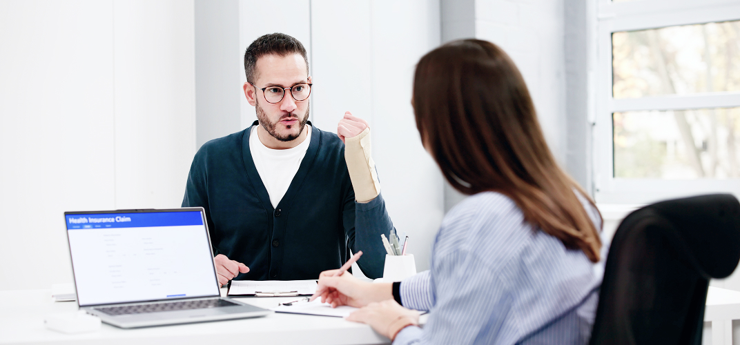 Man with a cast on his arm explains something to a woman across a desk in a bright office, laptop open nearby