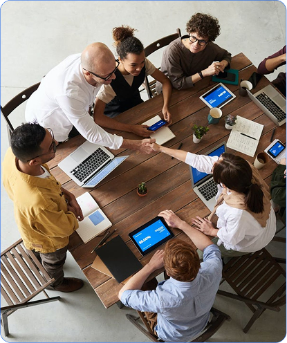 Group of diverse colleagues around a table, shaking hands over tablets and laptops in a collaborative meeting.