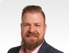 Head-and-shoulders portrait of a smiling man with a beard, wearing a dark blazer and pink shirt against a light background.