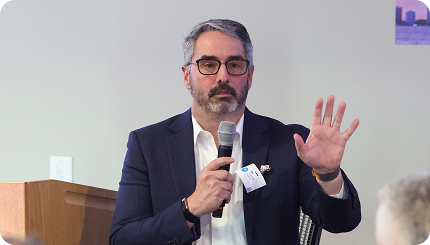 Man in a dark suit speaks into a handheld microphone while gesturing with his right hand at a conference/panel event.
