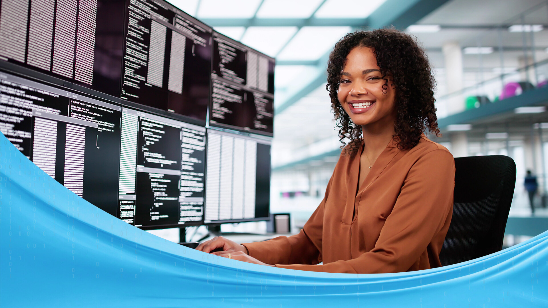 Smiling woman seated at a multi-monitor coding workstation in a bright office