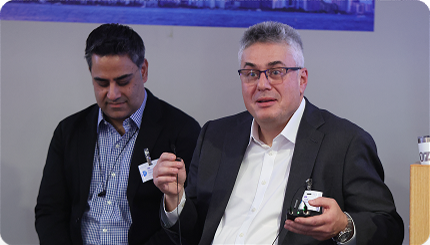 Two professionals on a conference panel; the man on the right speaks into a microphone while holding a small device, the other man looks down.