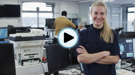 Video thumbnail: smiling woman in a navy blouse stands with arms crossed in a busy computer lab, with printers and monitors around; play button overlay indicates a video.