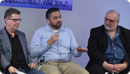 Three men sit on a panel; the middle man speaks and gestures with his hand while the others listen in a blue-lit room with a cityscape backdrop.