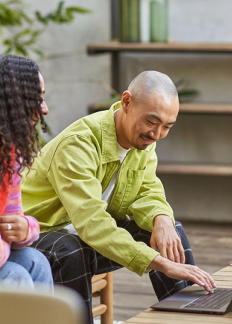 Two people sit together at a wooden table outdoors, the man in a lime‑green shirt typing on a laptop while a woman with braided hair sits nearby and watches.