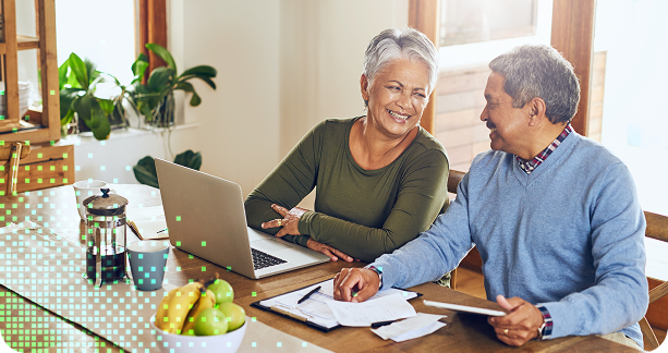 An elderly couple sits at a kitchen table with a laptop, papers, and a fruit bowl, smiling as they work together.